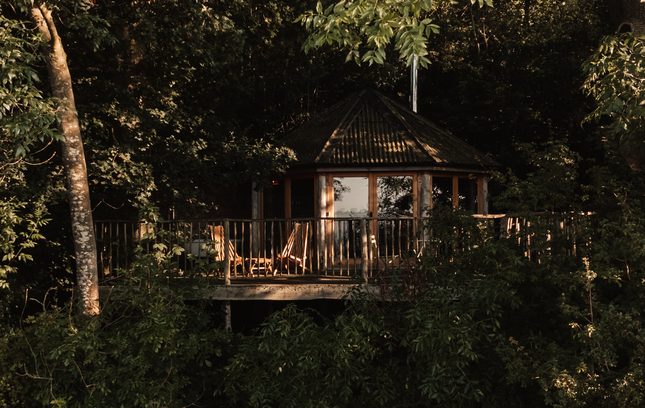 Treehouse surrounded by dense trees, with wooden deck and chairs.